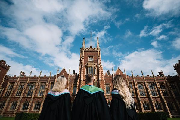 Three students in graduation gowns look up at the Lanyon