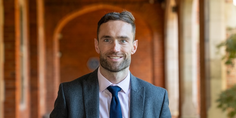 Portrait of Matthew Jackson in the Cloisters. Light blue suit, dark tie, young man with dark hair and beard.