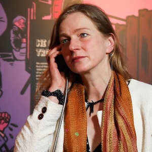 Portrait of Franziska Schroeder, with long hair and scarf, holding a telephone, with a colourful backdrop