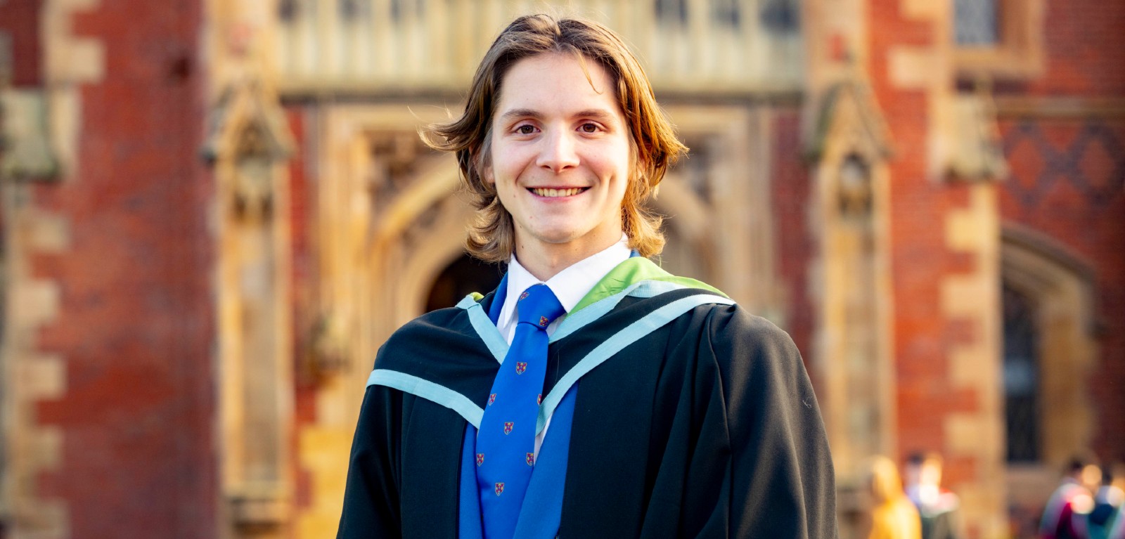 A male graduate stands in front of a university, smiling towards the camera