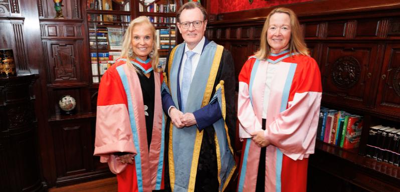 Two women and a man dressed in academic robes stand in a study room looking at the camera