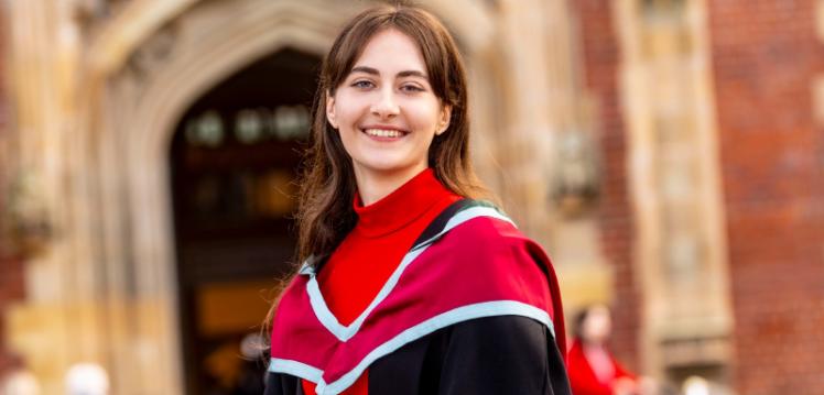 A female graduate stands in front of a university, smiling towards the camera