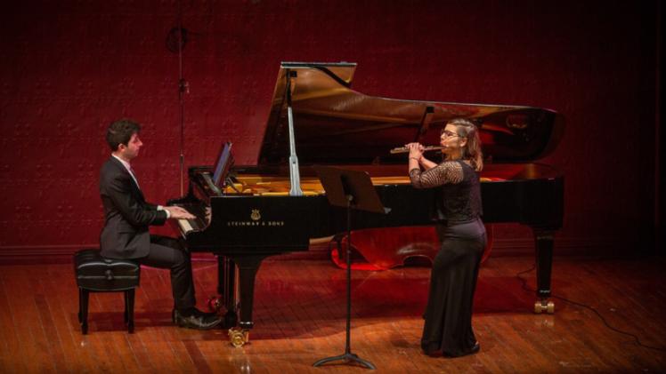 Person sitting at and playing a piano alongside a person standing and playing a flute.