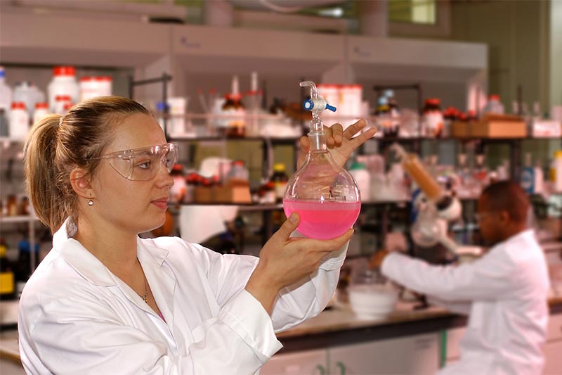 female student in lab coat examining a beaker filled with a pink liquid