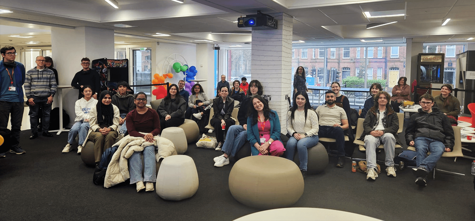 A group of people gathered in a modern indoor space with large windows, colourful balloons, and casual seating. The attendees are smiling and looking at the camera, seated on beanbags and chairs, with some standing in the background.