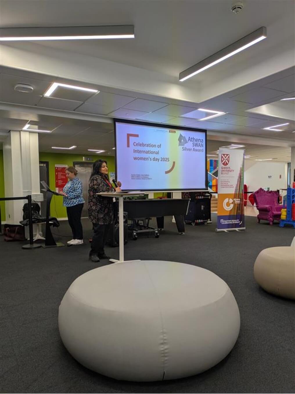 A speaker stands at a podium addressing an audience during a 'Celebration of International Women's Day 2025' event. A large screen behind her displays the event title along with the Athena SWAN Silver Award logo. A banner for Queen’s University Belfast and the Centre for Secure Information Technologies (CSIT) is also visible.