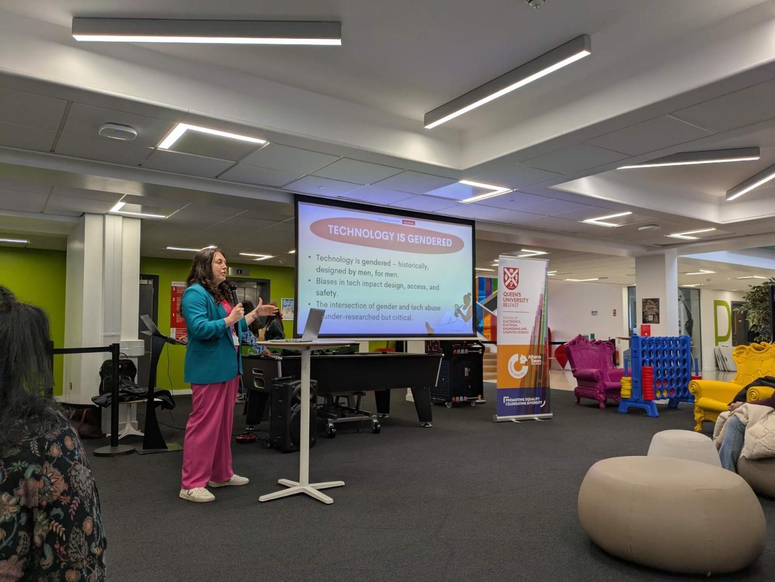 A speaker stands at a podium, behind her, a large screen displays a slide titled 'Technology is Gendered,' discussing historical biases in tech design and their impact on access and safety. A Queen’s University Belfast and Athena SWAN banner is visible to the right.