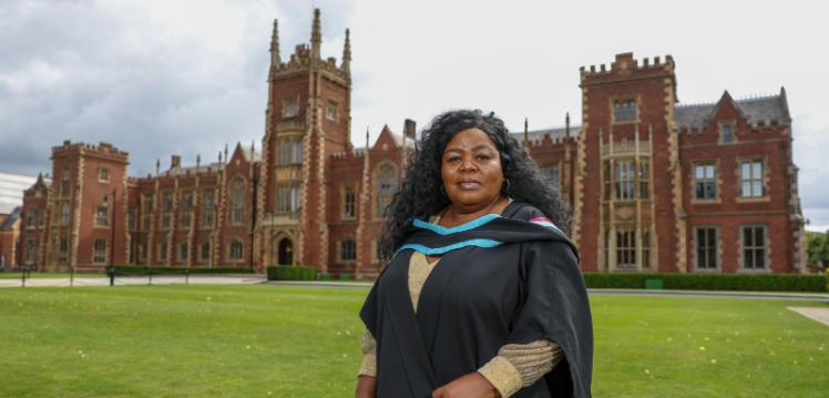 A woman with long black hair and wearing graduation robes, standing in front of the Lanyon building, a red-bricked building.