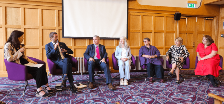 A panel of six people sitting in a wood-paneled room, engaged in a discussion during the 'Reflections: A 25-year Journey in Gender Equality’ event.