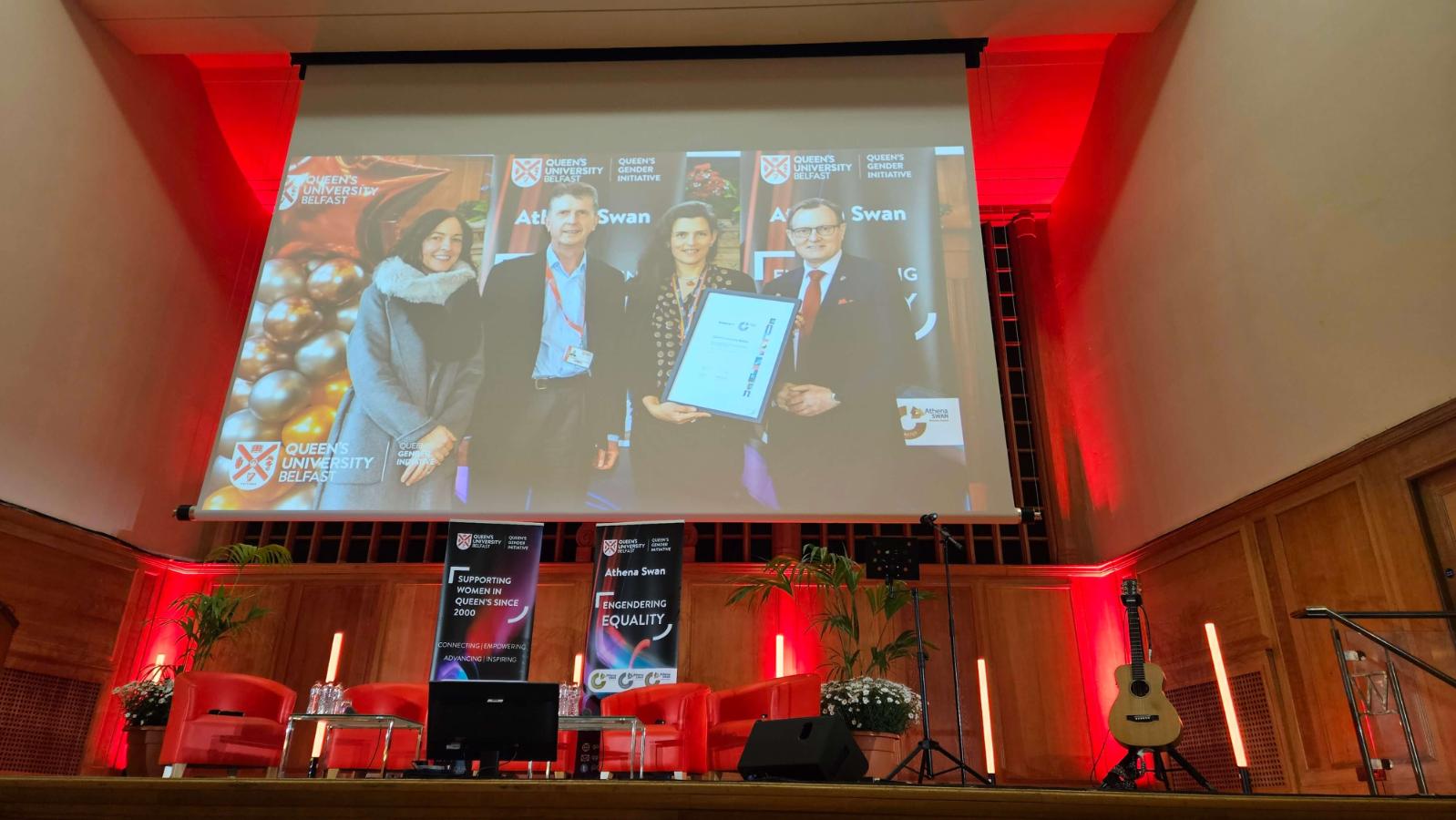 A stage set for an event with red chairs and a guitar, featuring a large projected image of people holding a framed certificate, with Queen's University Belfast and Athena SWAN branding in the background.