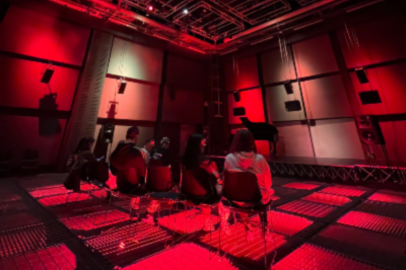 Group of people sitting in rows of chairs listening to music in a sound studio lit with red lights