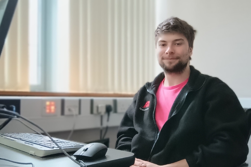 A young man sitting at a desk in a well-lit room with blinds covering the windows. He is wearing a black fleece jacket with a red logo and a pink shirt underneath, smiling softly at the camera. A computer keyboard and mouse are on the desk in front of him, with cables and electrical outlets visible in the background.