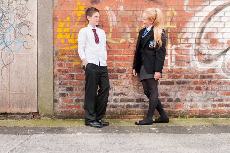 two teenagers in different school uniforms in front of a graffittee sprayed brick wall