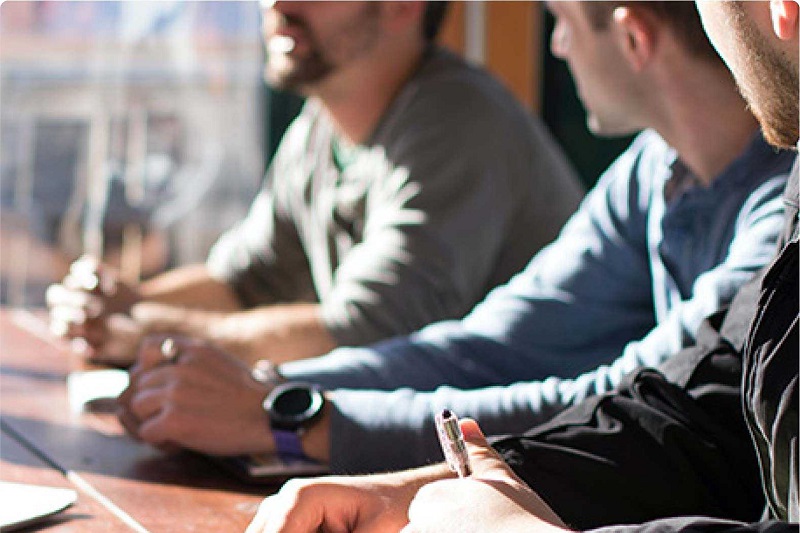 three seated men meeting at a table, chatting and taking notes