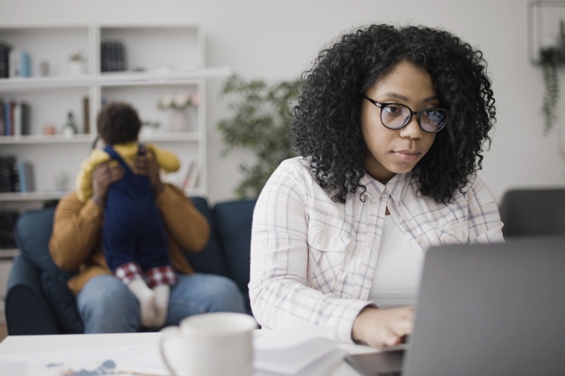 mother/woman working on laptop with father/partner playing with young child in background