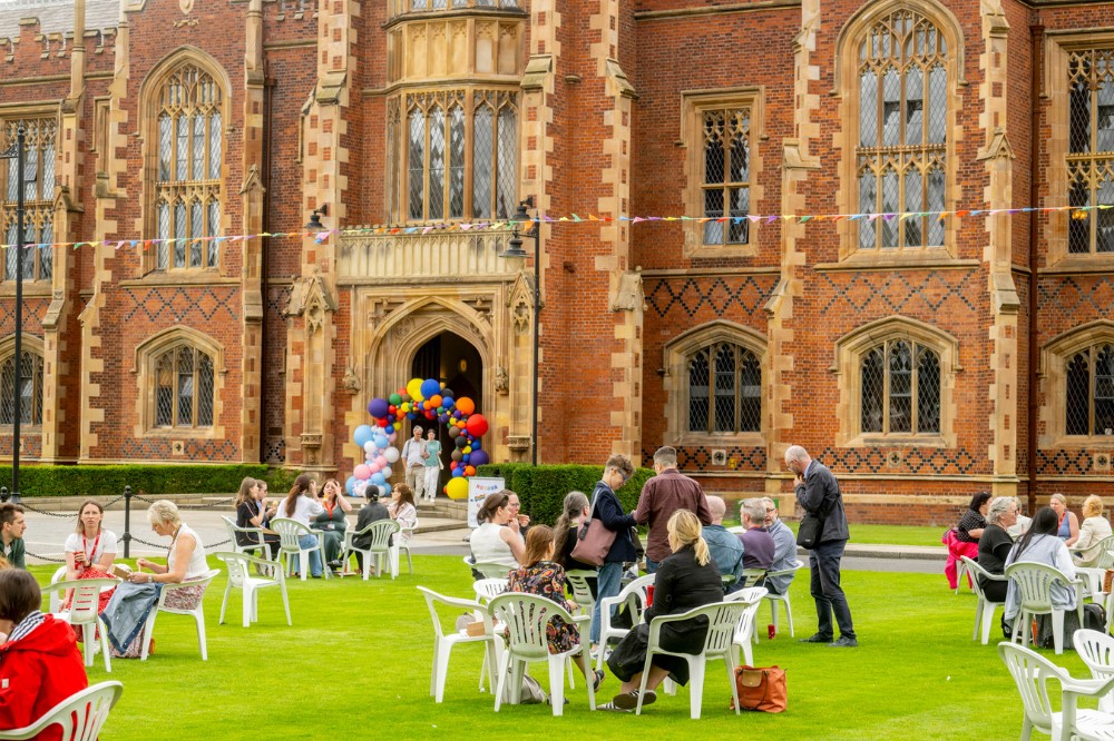 staff and students seated on the Queen's University Belfast front lawn at Queen's Pride Picnic 2024, with food trucks to the side and coloured bunting and balloons in sight